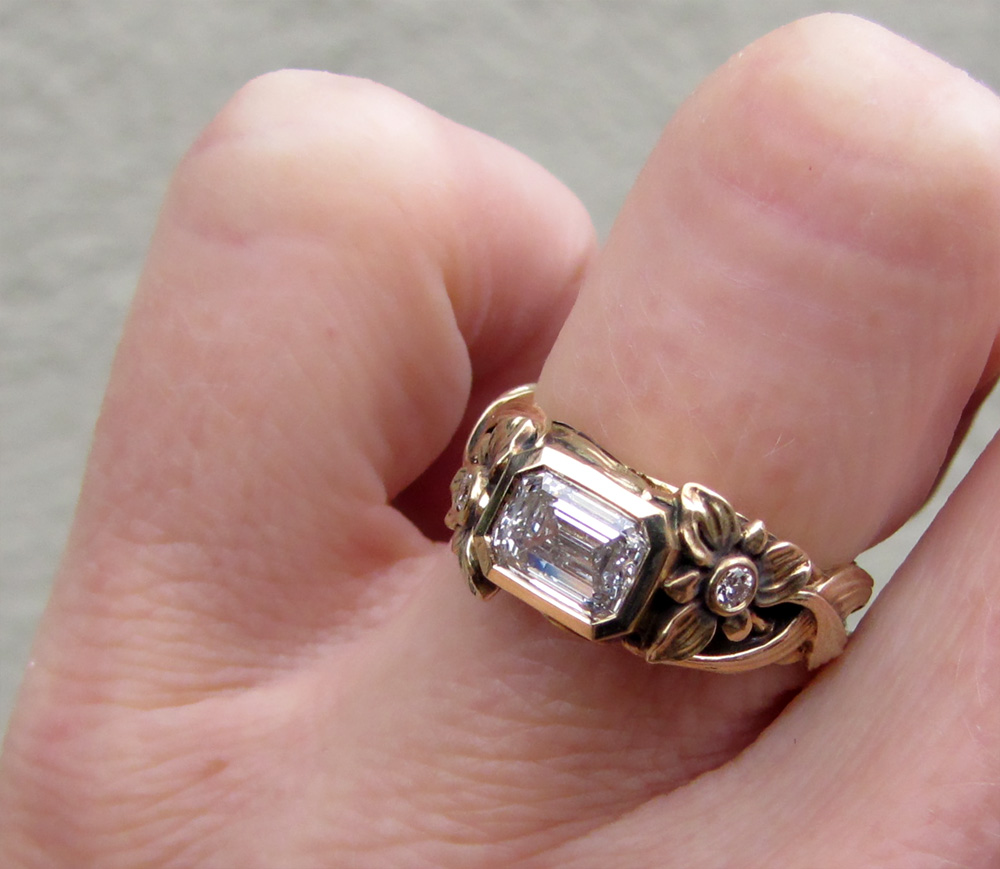 Custom-designed & hand-carved solitaire with 1ct emerald cut diamond. Lily flower, leaves, bulb motif.