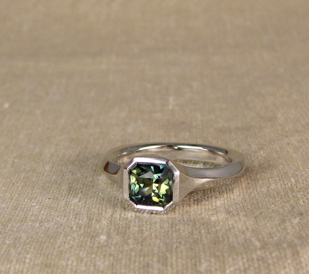 Custom hand-carved geometric solitaire for radiant-cut blue-green parti-sapphire. Platinum.
